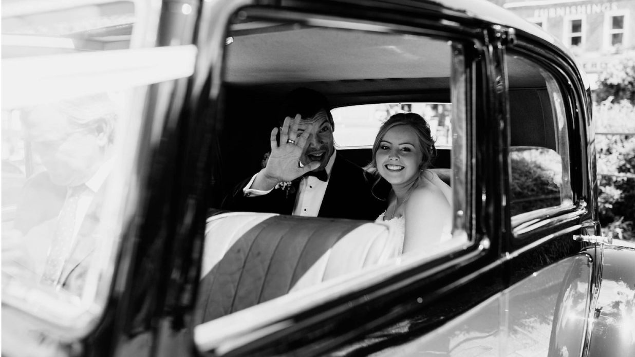 Black and White photo taken through the window of the vintage Rolls-Royce with Bride smiling and groom waving