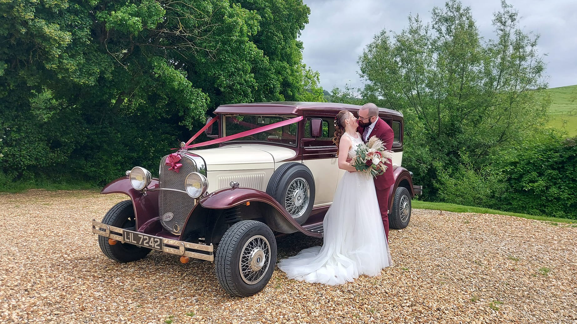Bramwith Limousine in Ivory and Burgundy dressed with white ribbons with Bride and Groom standing in front of the vehicle kissing.