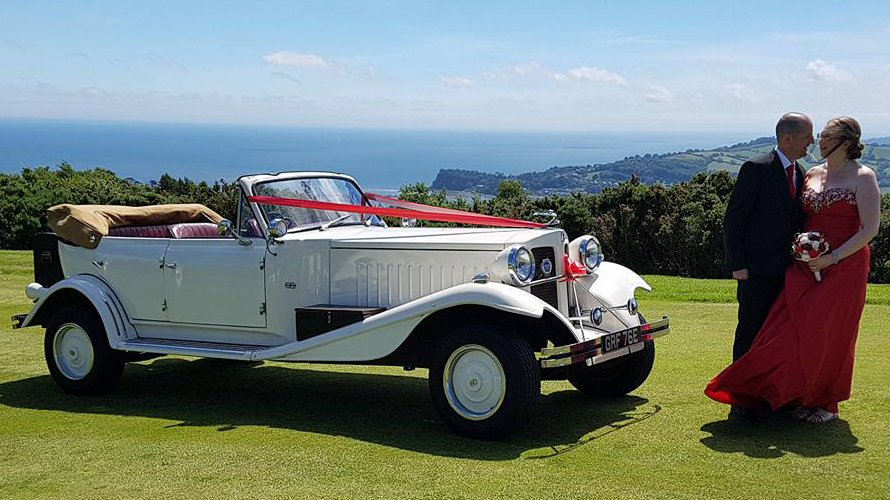 Beauford 4 Door Convertible on top of cliff decorated with burgundy ribbons with bride and groom standing in front of the vehicle