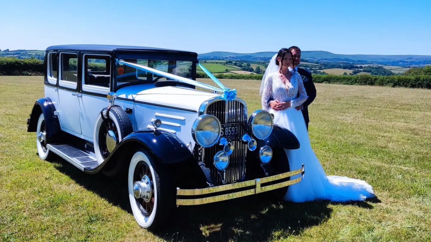 Black and Ivory Franklin dressed with light blue ribbons with Bride and Groom holding each other next to the car. View of the sea in the background