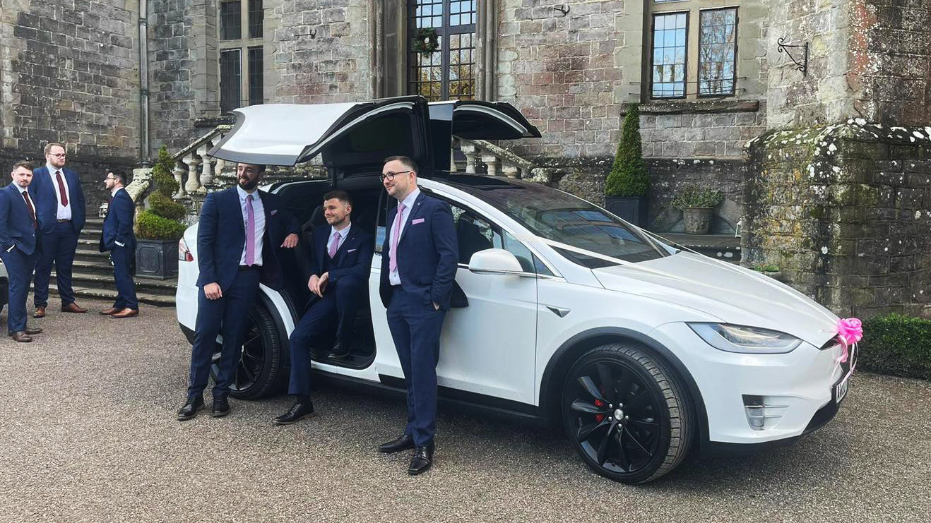 Tesla X in white decorated with white ribbon across the bonnet and groomsmen wearing navy blue suits standing around the car posing for their wedding photographer.