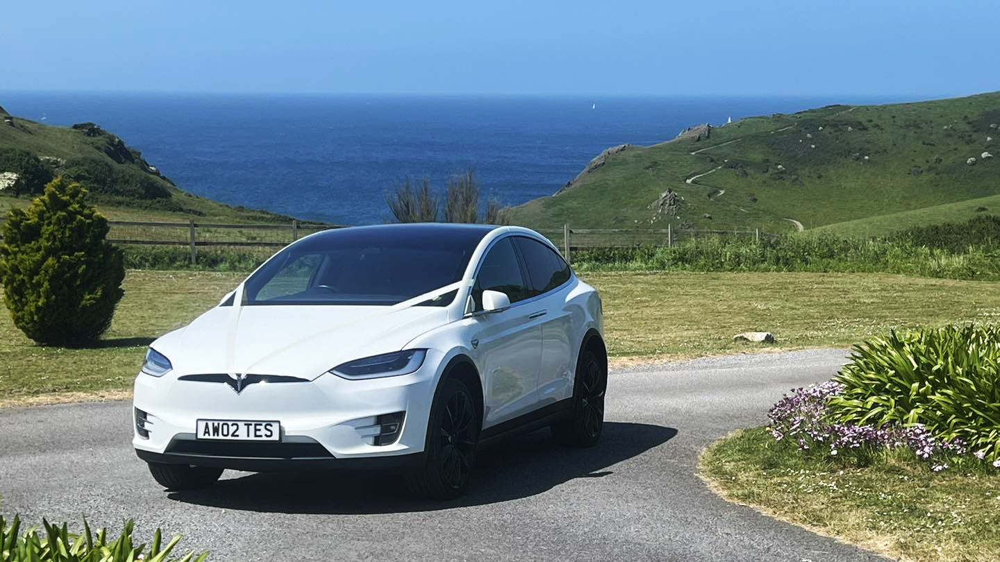 Modern White Electric Tesla X parked in the middle of the road on the Cornwall sea side with view of sea in the background