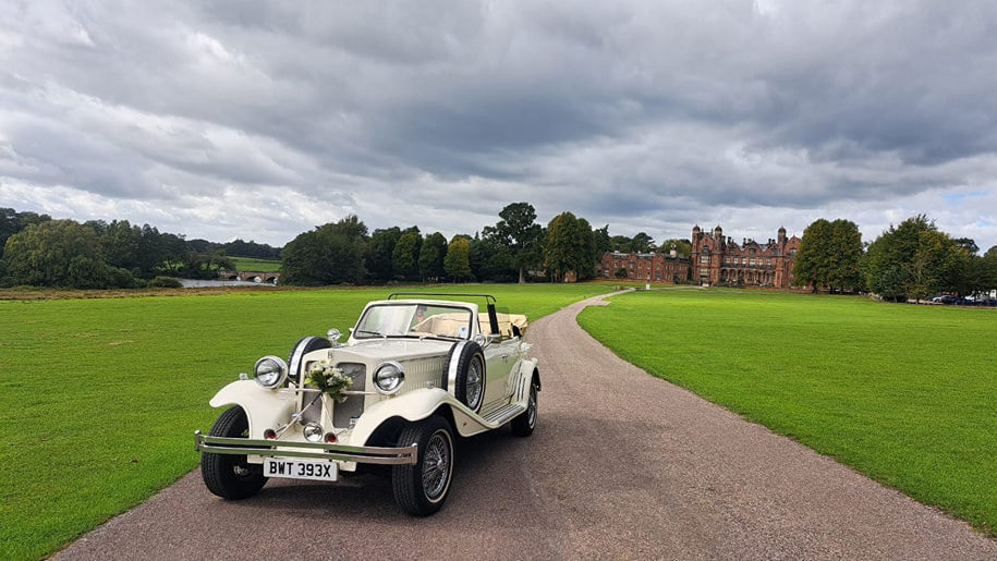 White Vintage Beauford Convertible with the roof down decorated with white ribbons and floral arrangement on front grill parked at the end of a long path with view of the wedding venue in the background