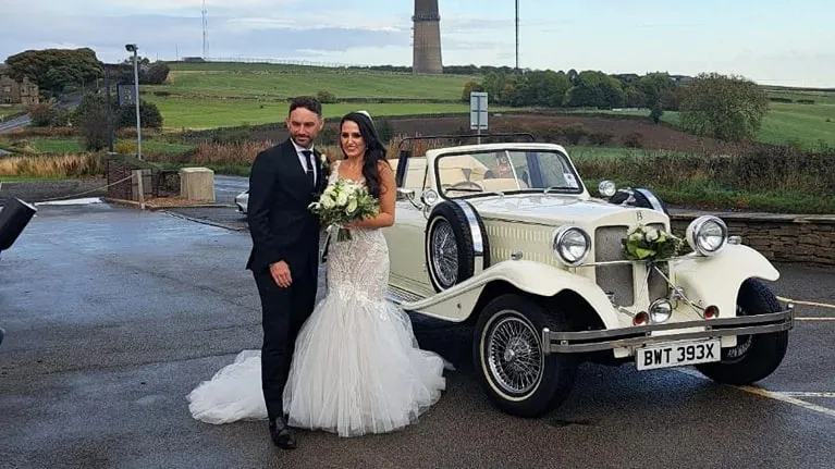 Ivory Vintage Beauford Convertible with roof down with Bride and Groom standing in front of the vehicle holding each others. Bride is wearing a white dress holding her bridal bouquet.