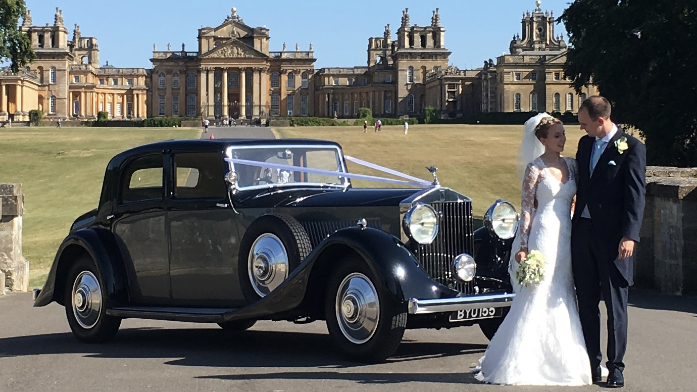 Rolls-Royce Phantom II Continental LWB decorated with white ribbon with bride and groom in front of the vehicle. Wedding Venue in the background