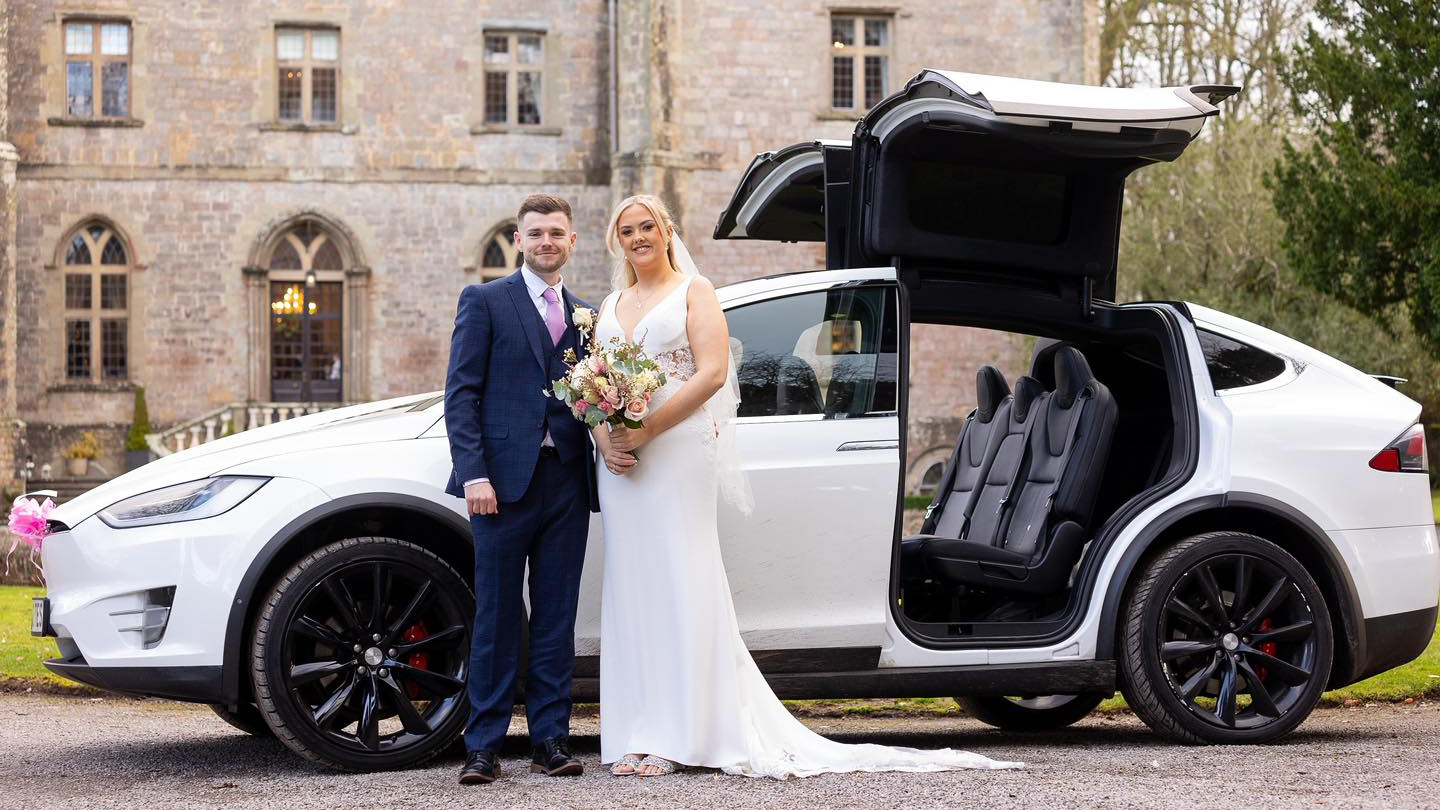 Bride and Groom stansing in front of a white Tesla X with Gull-wings open in front of a wedding venue