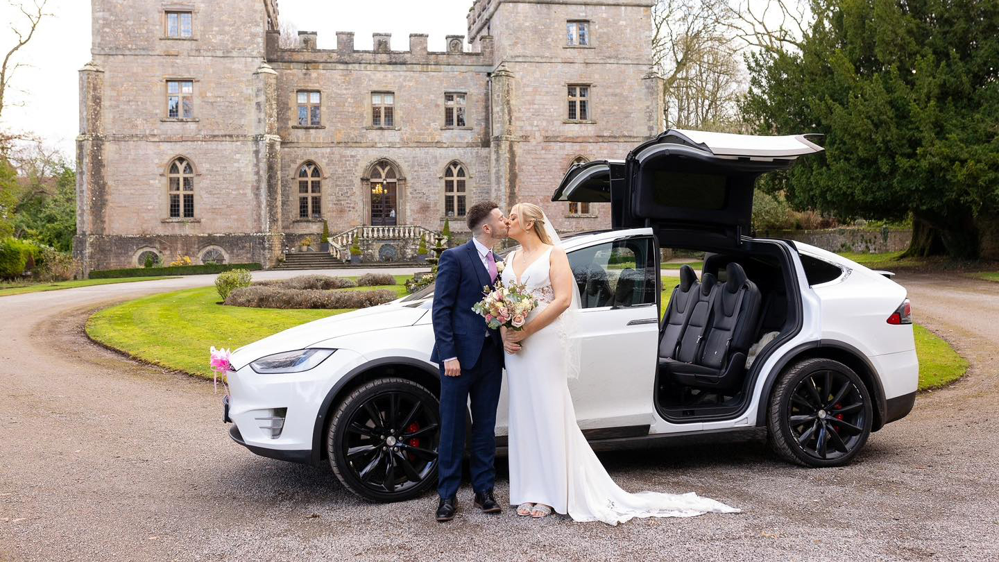 Tesla X in white parked in front of castle style wedding venue with its gull wings open. Bride and Groom are standing in front of the vehicle kissing.