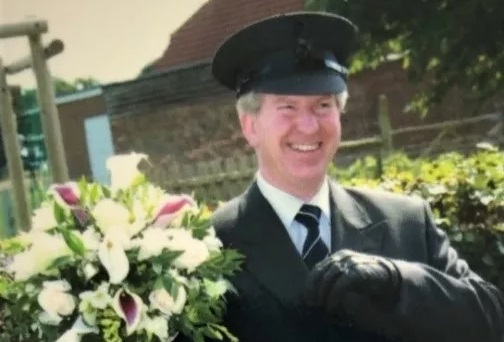 Wedding car chauffeur wearing a dark grey suit, chauffeur's hat and gloves holding the bride's bridal bouquet