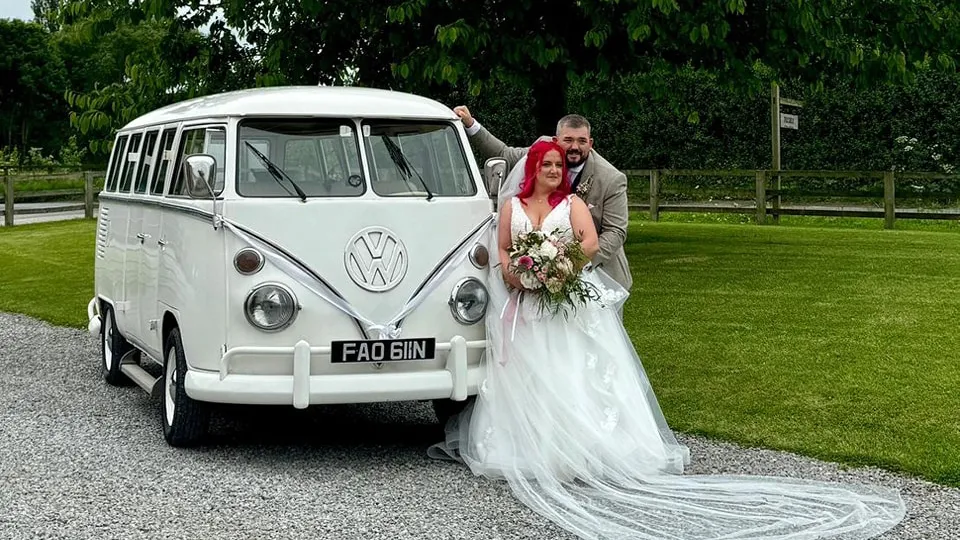 Classic VW Campervan dressed with white ribbons with Bride and Groom standing in front of the vehicle posing for their photographer. Vehicle is parked in the garden of a local wedding venue in Lancaster.