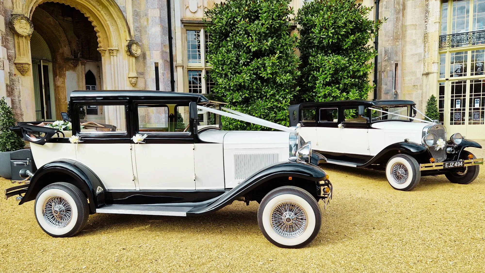 Two matching vintage wedding cars decorated with matching white ribbons. Vehicles are parked at the front entrance of a local wedding venue in Swanage.