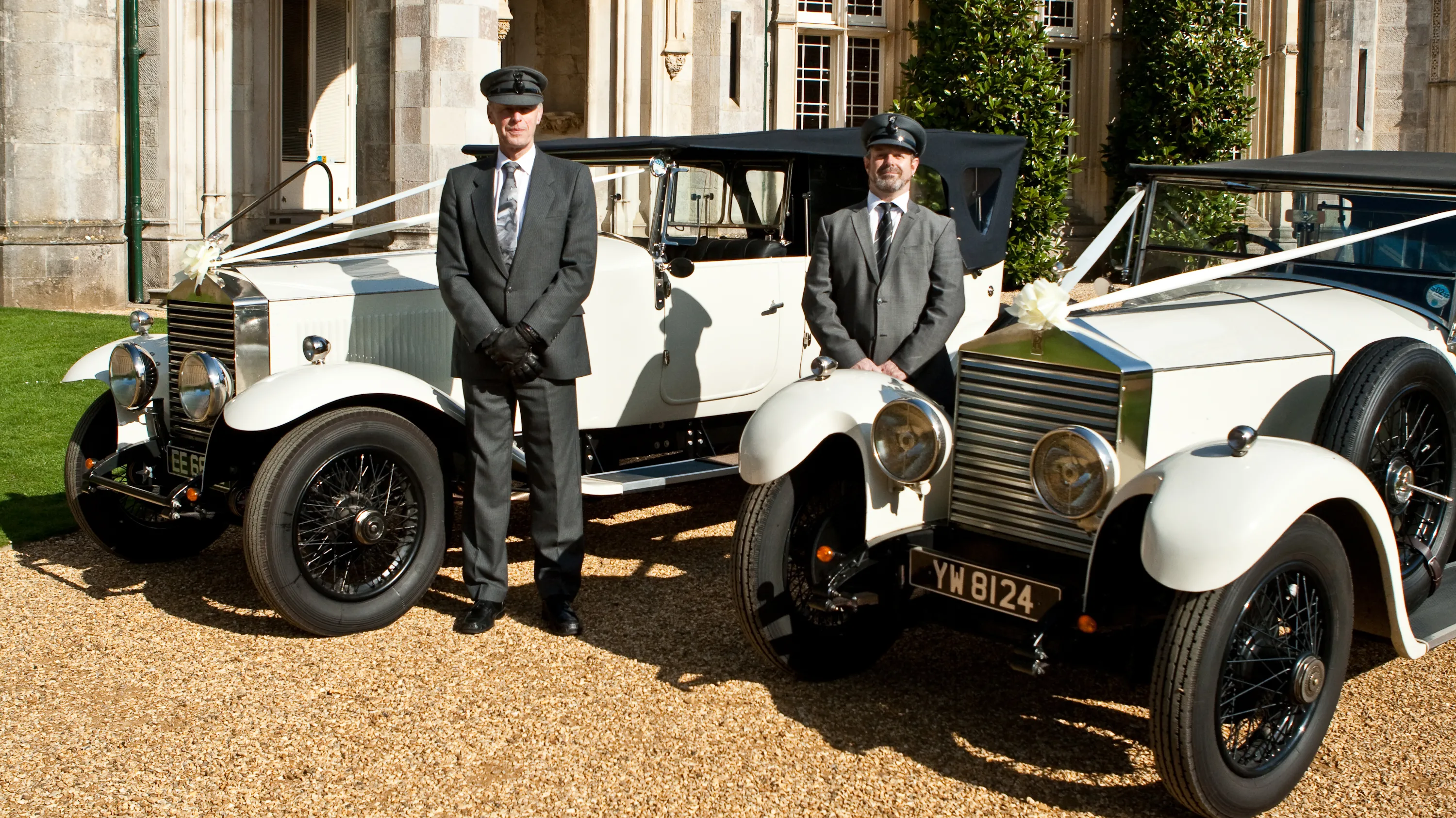 Two vintage wedding cars decorated with white ribbons and bows at a local Bournemouth wedding. Both vehicles are parked side-by-side with their chauffeurs next to them.