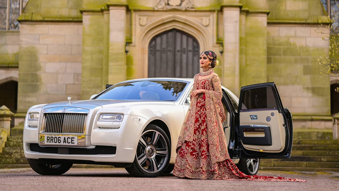 white Rolls-Royce Ghost with its forward facing door open with Asian Bride in a traditional wedding dress standing by the vehicle. A popular Lancashire wedding venue can be seen in background.
