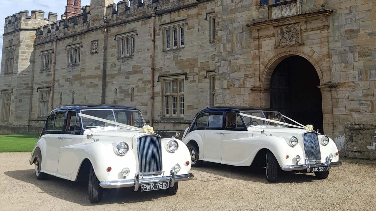 Two Matching Black & White 7-seater Austin Princess Limousines decorated with matching ribbons and bows with castle style wedding venue in the background.