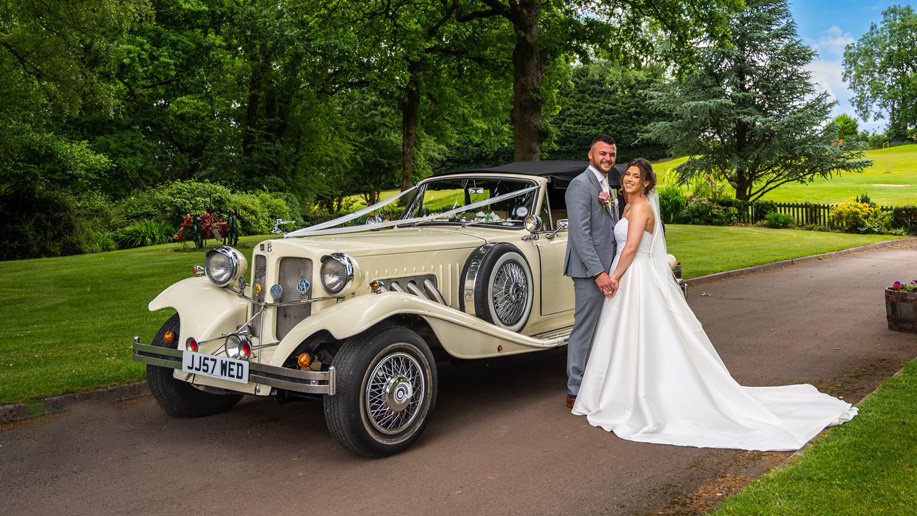 Ivory Convertible Beauford with black soft-top up decorated with white ribbons with bride and groom holding hands and smiling.