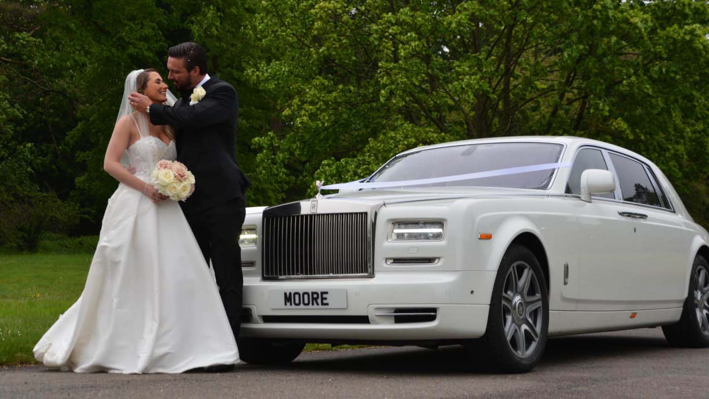 White Rolls-Royce Phantom with traditional white ribbons across the bonnet. Bride and Groom are standing in front of the vehicle kissing.