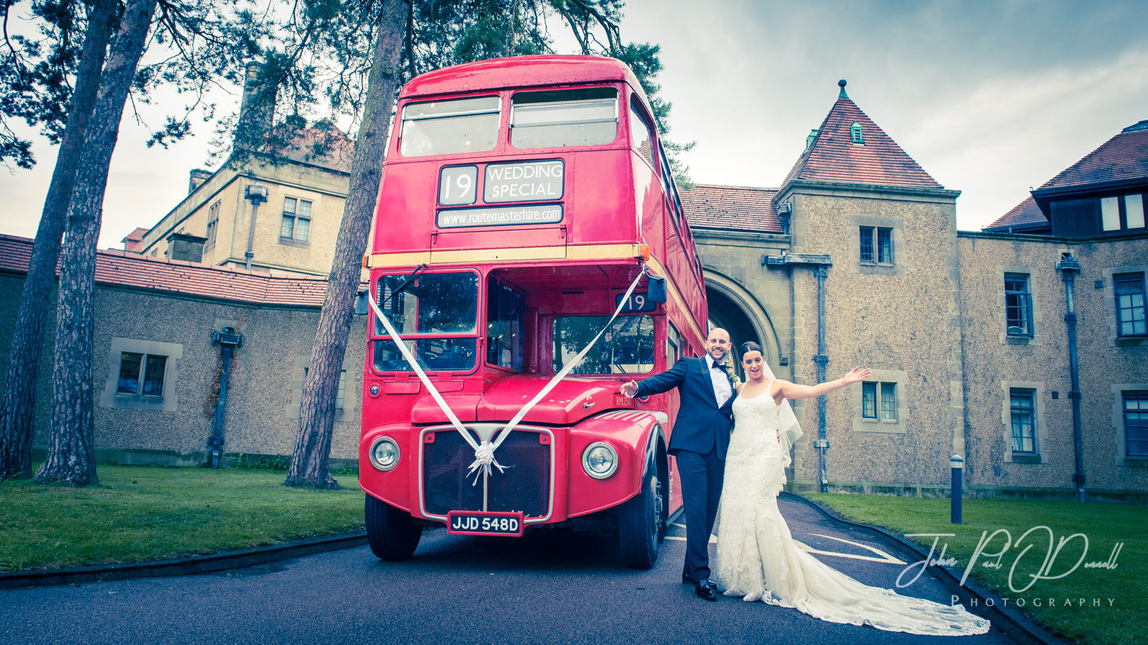 Double Decker Red Routemaster Bus with white ribbons. Bride and Groom are standing in front of the bus smiling