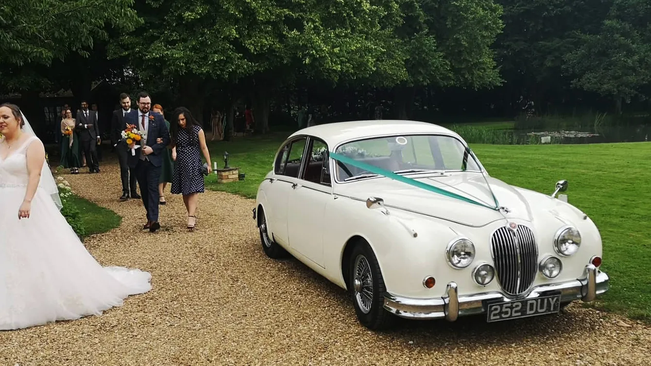 Classic Jaguar mark 2 decorated with V-Shape turquoise ribbons. Bride and wedding guests are walking passed the vehicle.