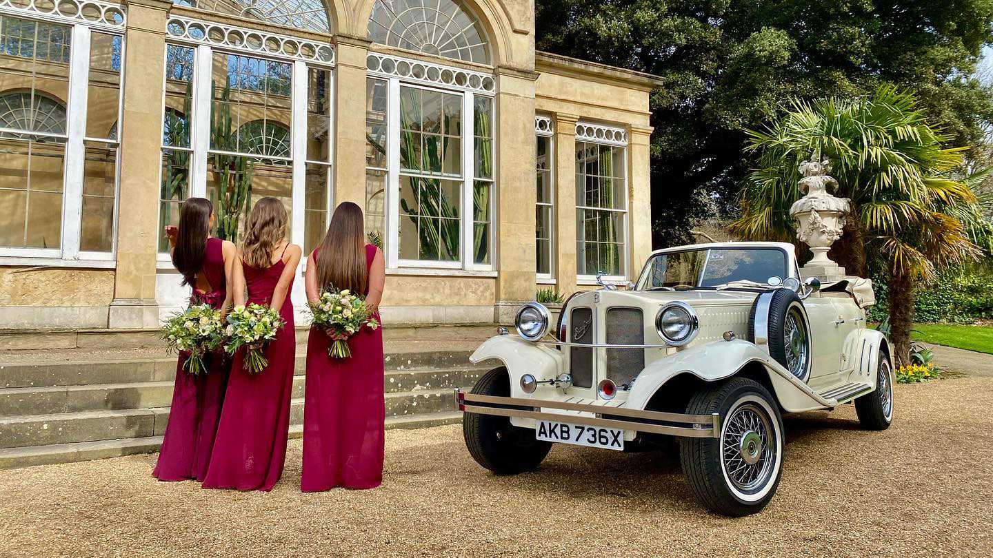 Ivory Beauford convertible with roof down parked in front of a wedding venue with three bridesmaids standing in front of the vehicle facing the vehicle in Ruby Red dresses holding their bridesmaids bouquet behind them.