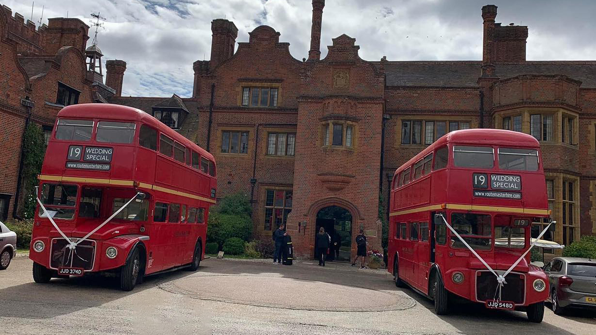 Two Routemaster buses parked side-by-side with matching white ribbons in front of a wedding venue