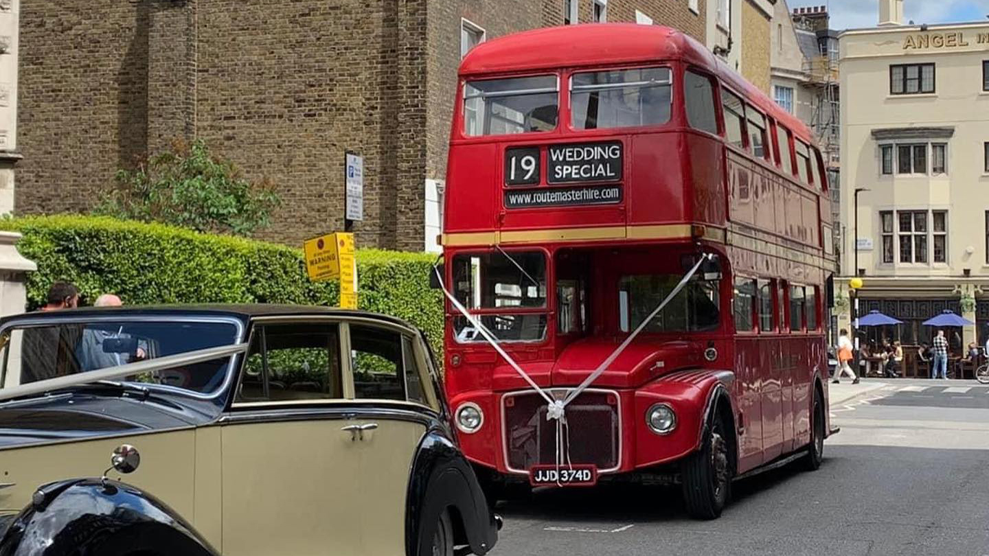 Front view of a Red Routemaster bus with white ribbons following a vintage car in the street of St Albans in Hertfordshire
