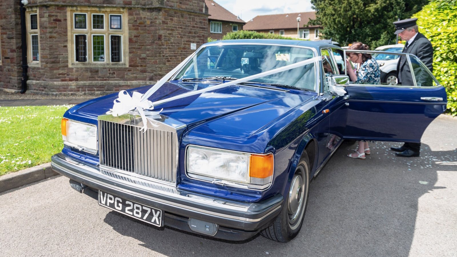 Blue Rolls-Royce Silver Spur decorated with White Ribbons and bows. Uniformed chauffeur is opening the rear door to let the bride out.