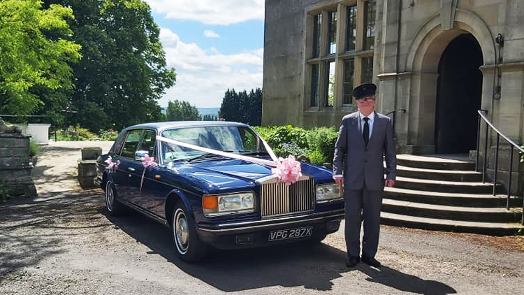 Blue Rolls-Royce Silver Spur with pale pink ribbon in fropnt of a wedding venue. Fully uniformed chauffeur standing in front of the vehicle in his grey suit and hat.