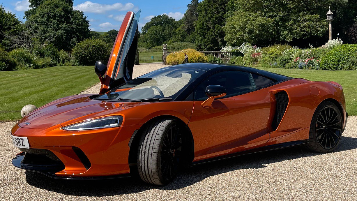 Burnt orange coloured McLaren GT in the garden of a wedding venue with one of its scissor door open