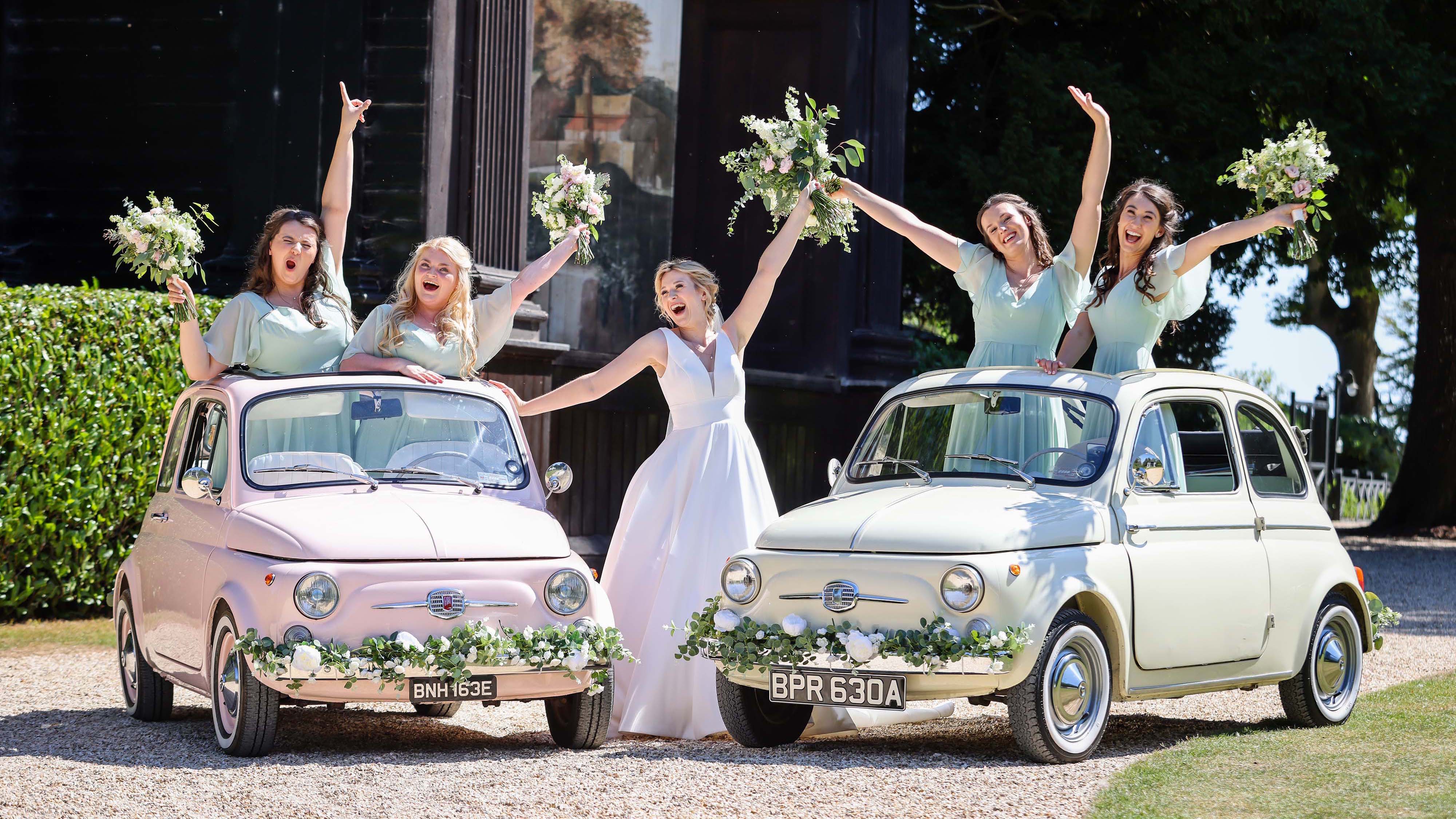 Two Classic Fiat 500 wedding cars in pale pink and blue with floral decoration on the front chrome bumper. Bride and her bridesmaids are standing with the vehicles cheering and smiling.