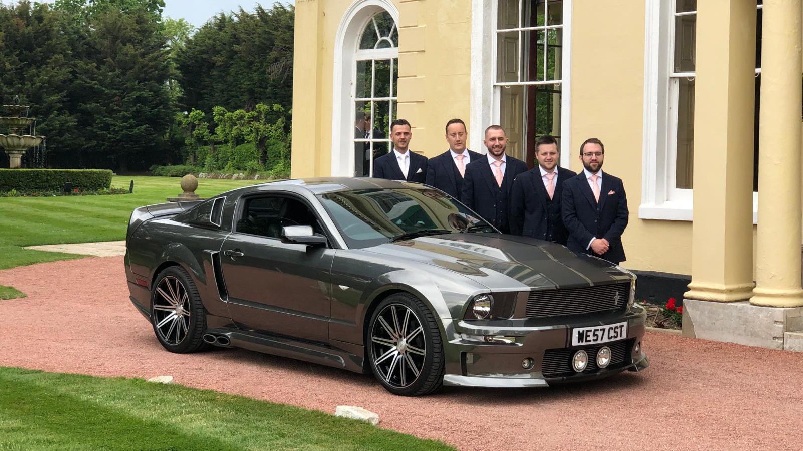 Grey Ford Mustang V8 parked in front of a wedding venue with Groom and his Bestmen standing behind the vehicle.