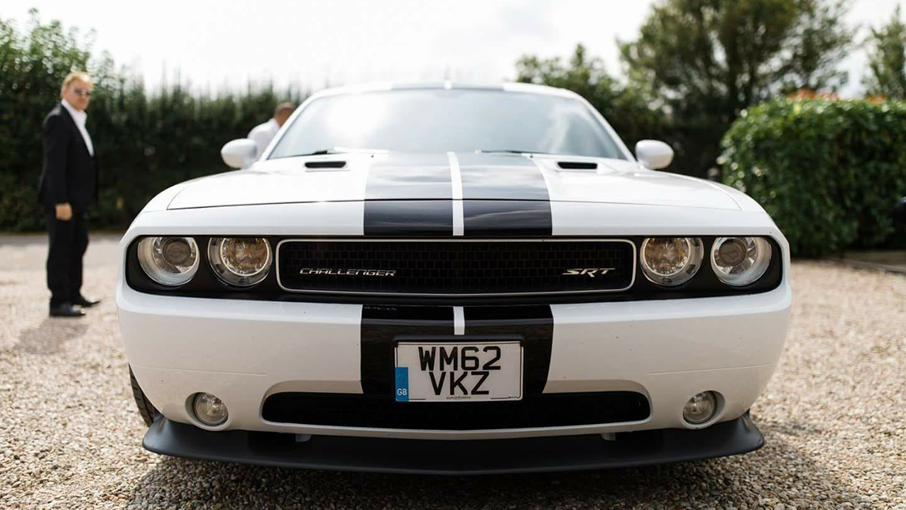 Front view of a white American Dodge Challenger with black stripes across front bonnet. Groom stands in the background on the left looking at the vehicle