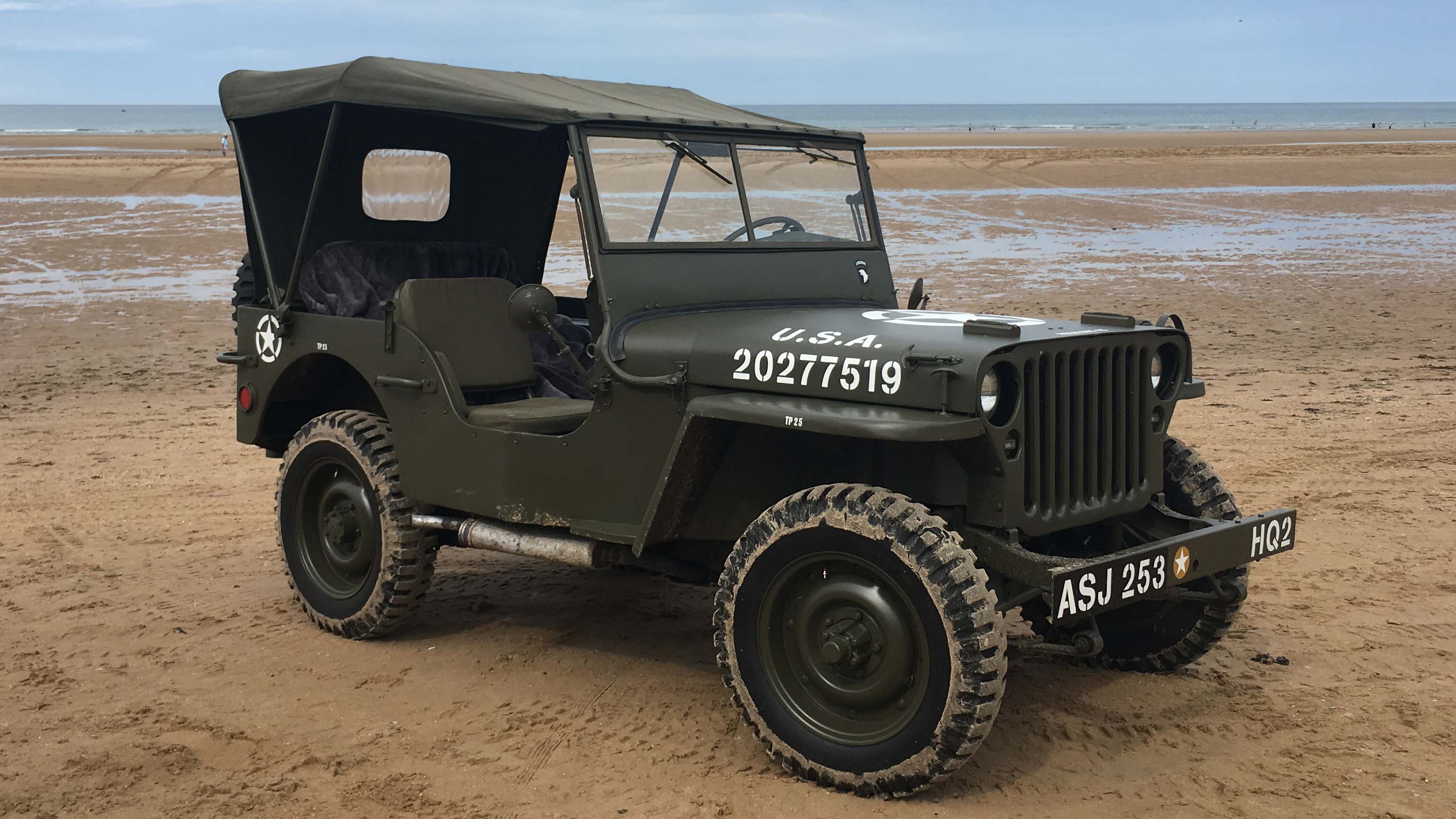 Khaki Green Military Jeep with its roof up on a sandy beack with sea in the background