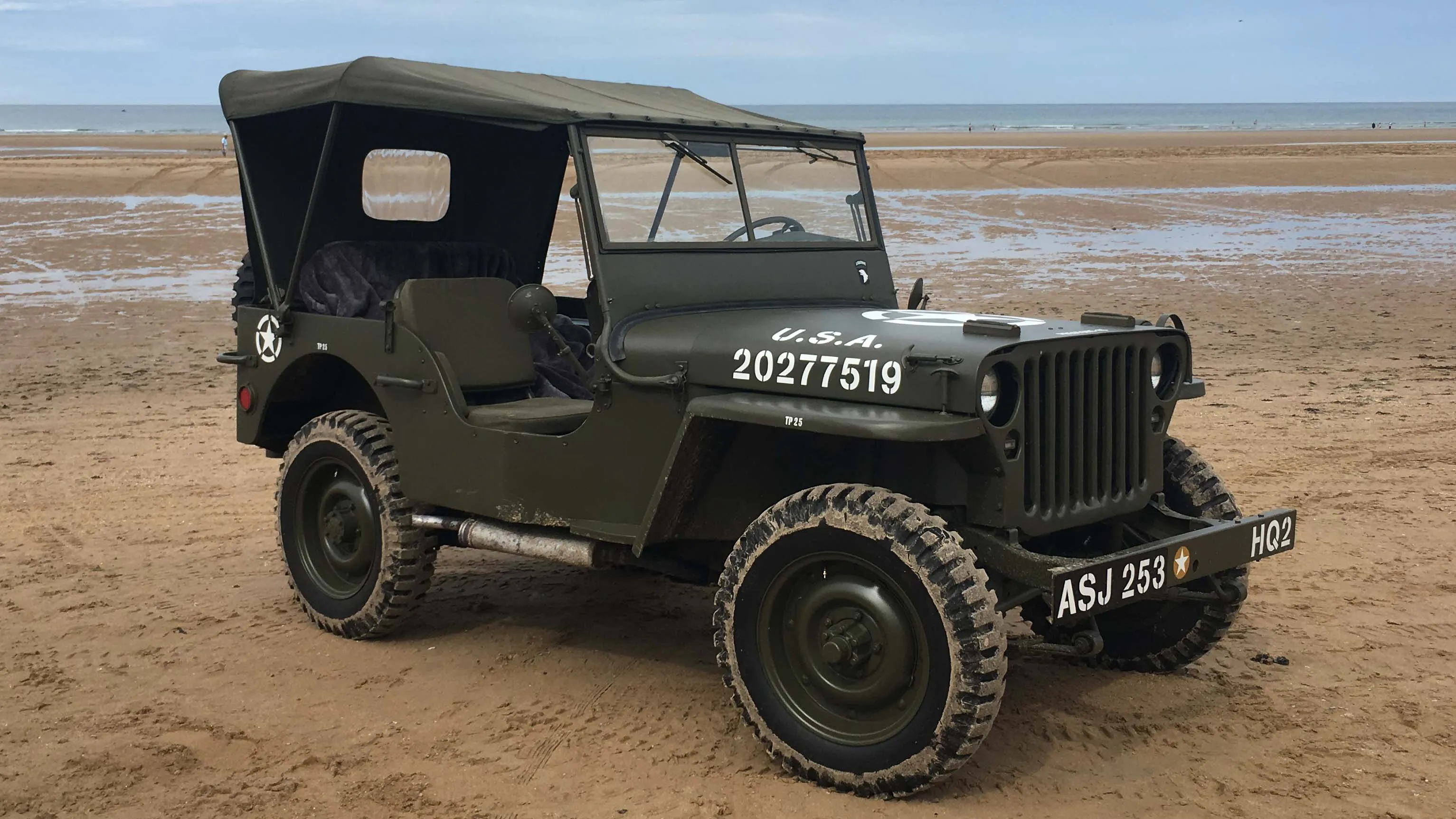 Khaki Green Military Jeep with its roof up on a sandy beack with sea in the background