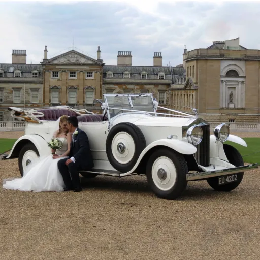 Vintage Rolls-Royce Convertible  ALT=White Vintage Rolls-Royce Convertible with roof down. Bride and Groom are seated on the skirt of the vehicle.