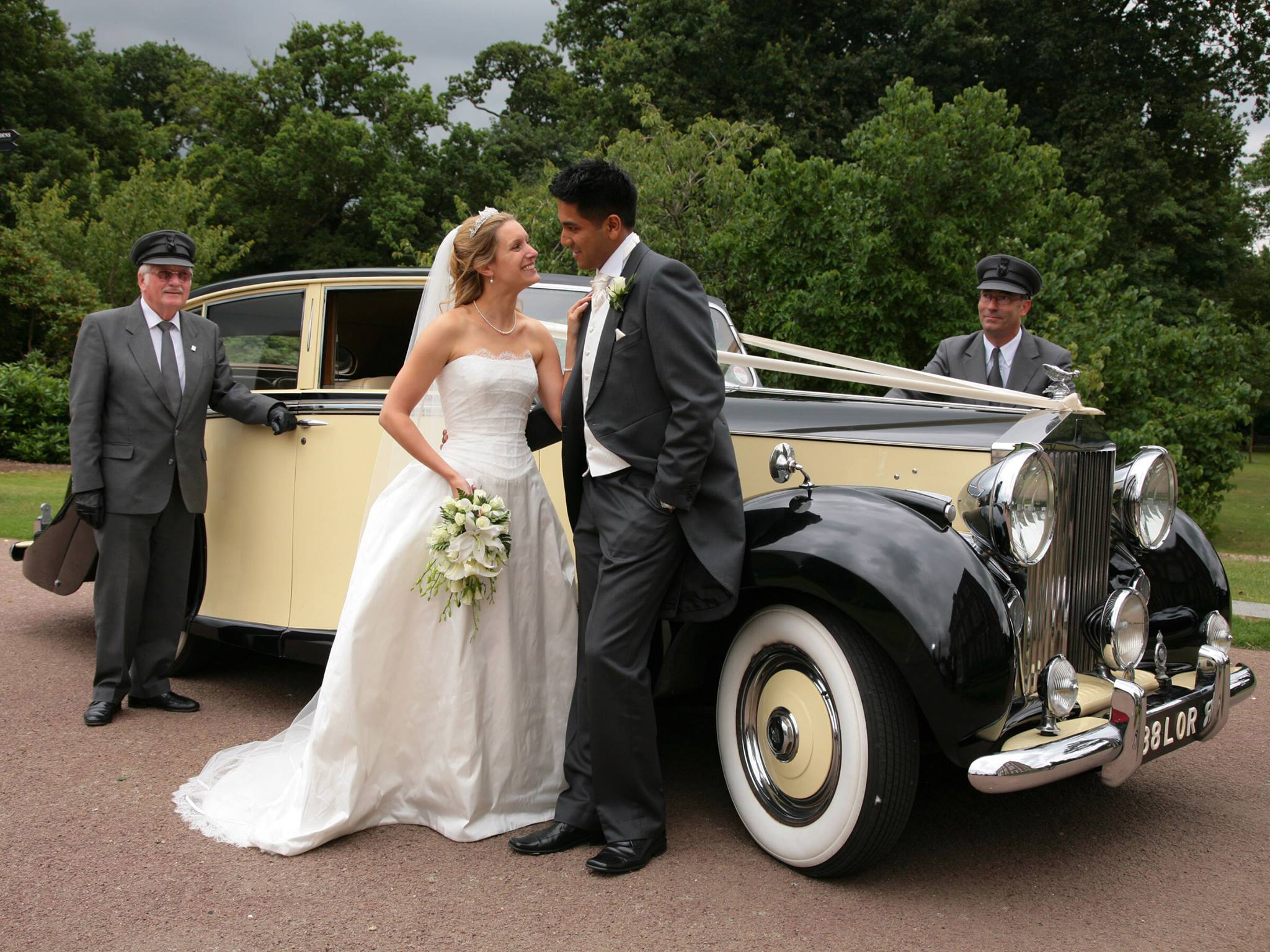 Two experienced chauffeurs wearing a grey uniformed with chauffeur’s hat standing next to a classic Rolls-Royce looking at smiling Bride and Groom.