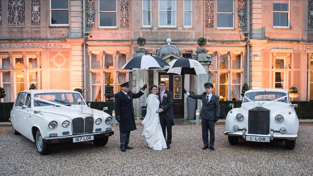 Two Full uniformed chauffeurs holding umbrellas over the Bride and Groom in the middle of two classic cars.