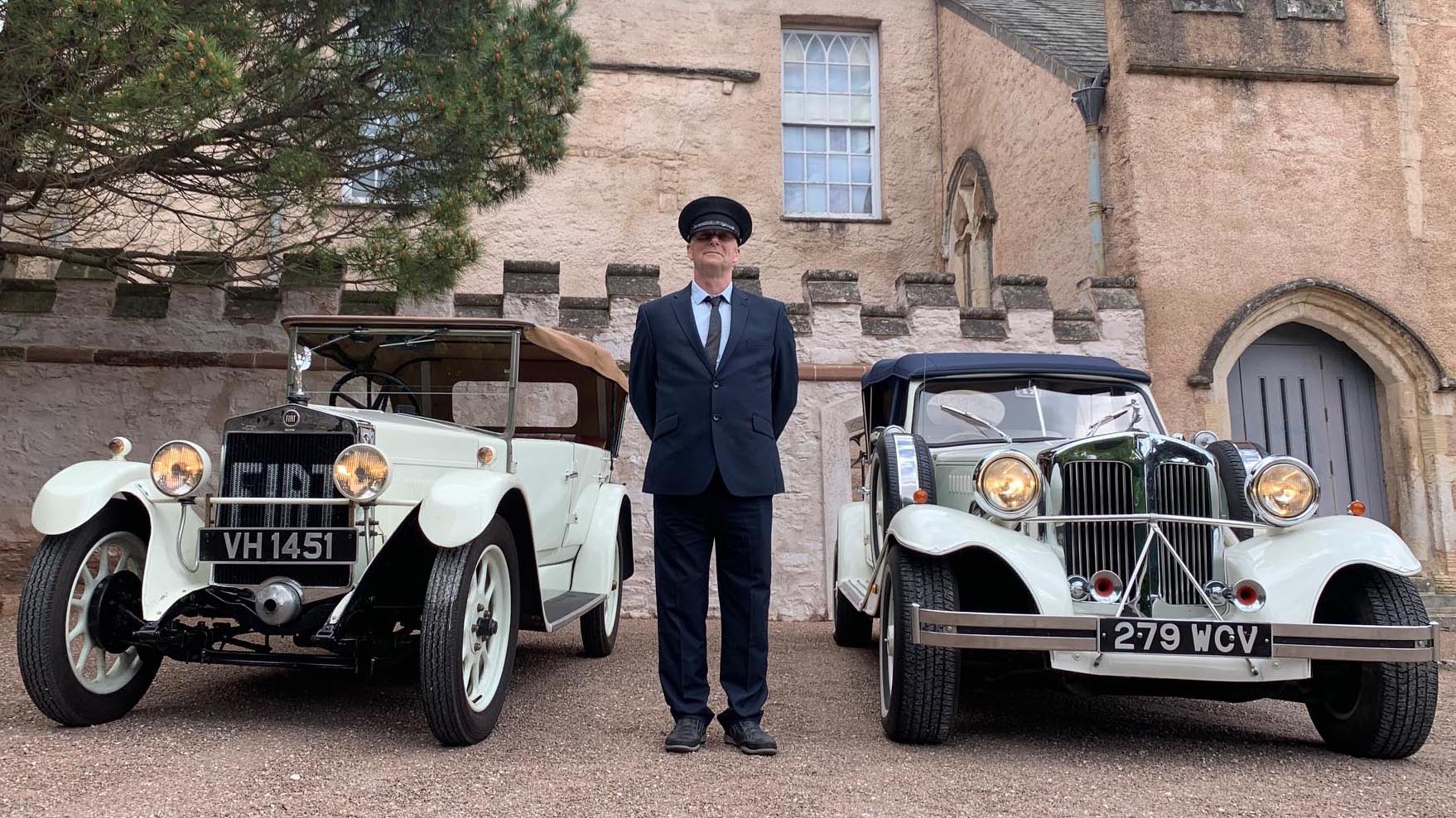 Fully uniformed chauffeur with hat standing in the middle of two white vintage cars.