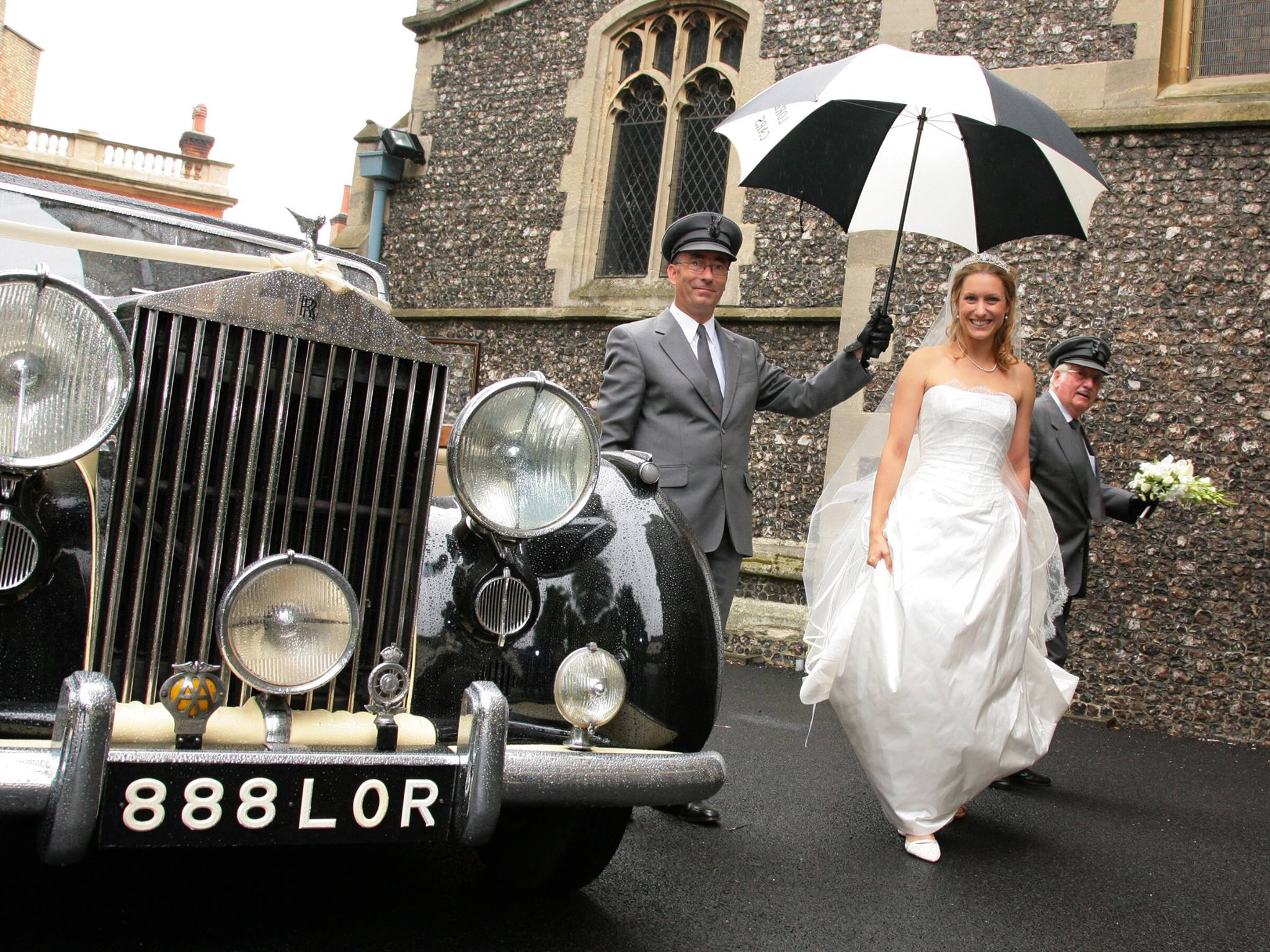 Wedding car chauffeur walking a smiling bride wearing a white dress to the church holding an umbrella in his hand.