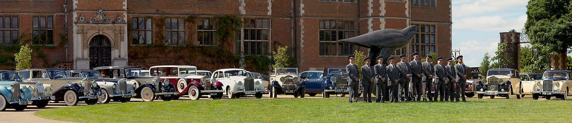 Chauffeurs wearing full grey uniform and hat in front of a selection of classic cars.