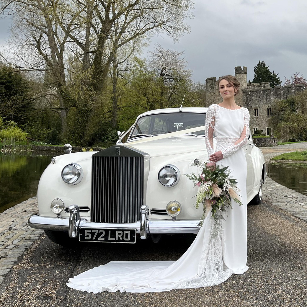 White Rolls-Royce Silver Cloud with Bride standing in front of the vehicle holding her bridal bouquet. Wedding venue can be seen in the background.