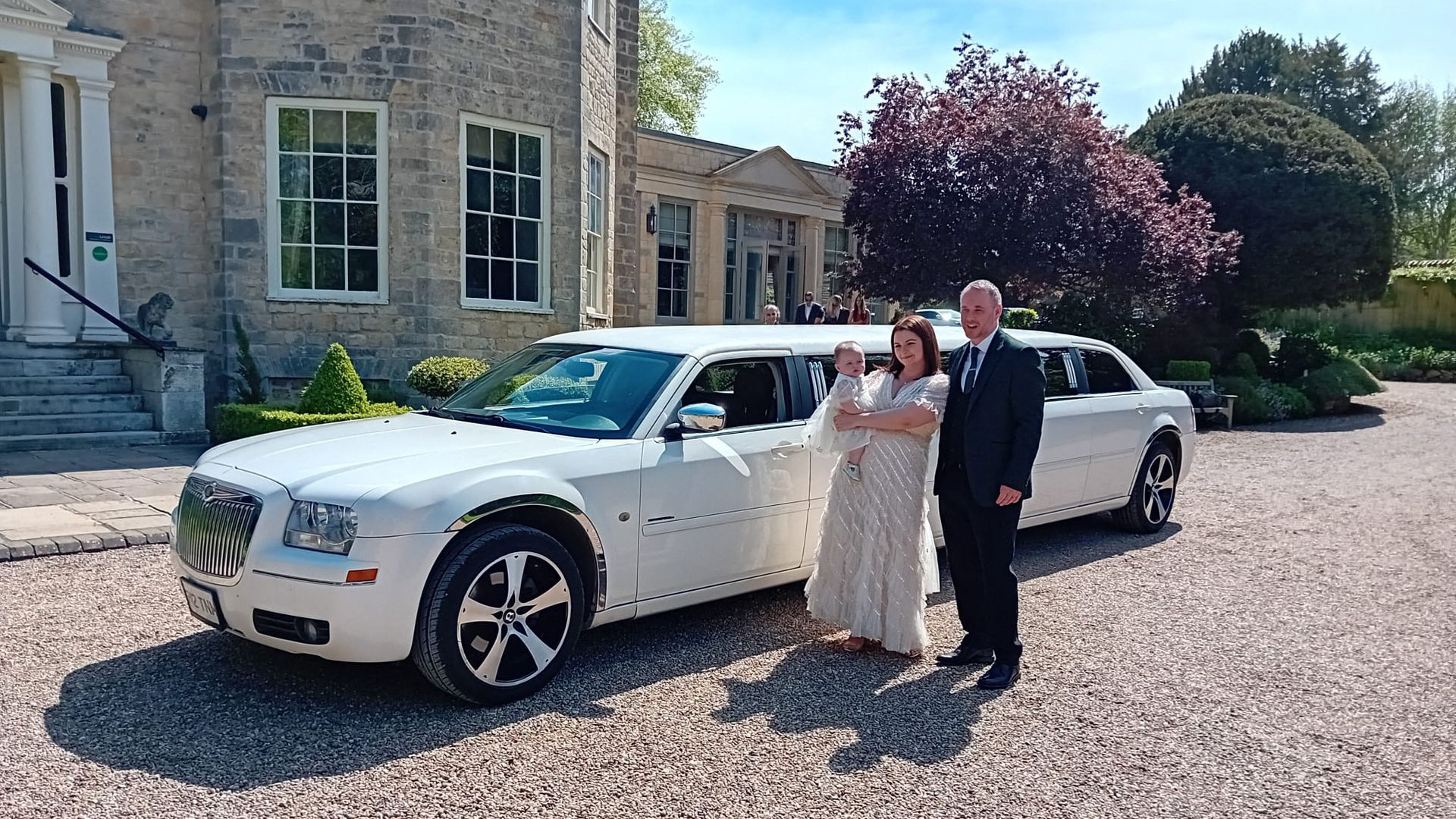 Chrysler 300c White Limo in front of a wedding venue with Bride and Groom standing by the vehicle. The Bride is holding her little infant baby in her arm.