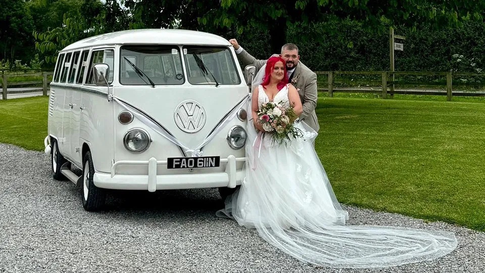 Classic VW splitscreen Campervan decorated with white ribbons at a local Sheffield wedding venue with Bride and Groom standing in front of the vehicle. Bride wears a white dress with bright red hairs and groom wears a light brown suit