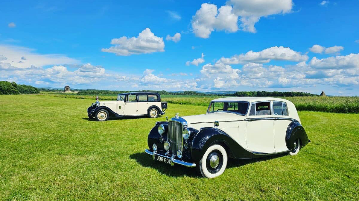 A Classic and Vintage Cars, in matching Black & Ivory colours, parked in a green field and a sunny blue sky