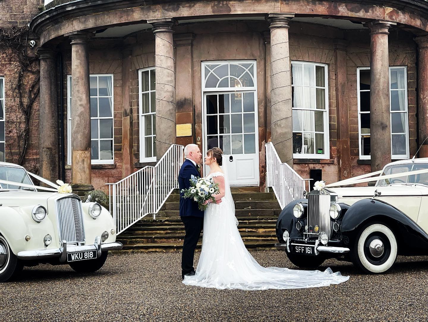 Bride and Groom facing each others in front of a venue near Sheffield with two classic wedding cars on either side of the couple. Bride is holding a large bouquet of flowers