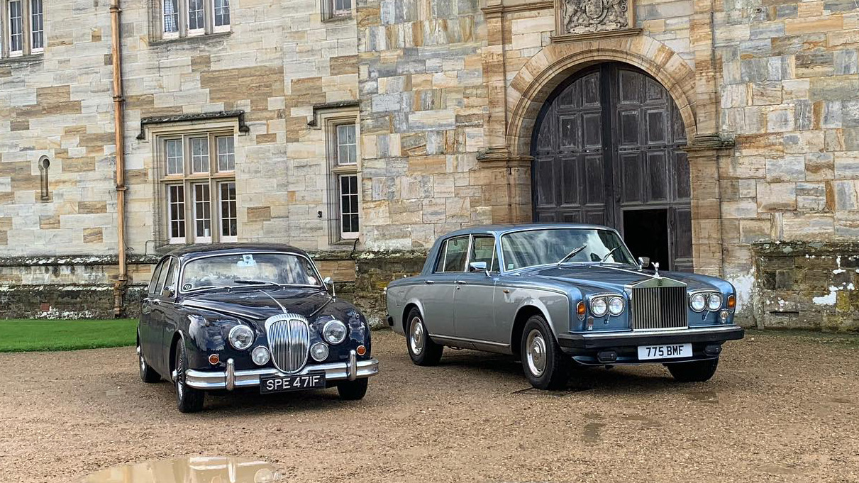 Two Wedding Cars decorated with matching white ribbons in front of a wedding venue. Vehicle on the left is a dark Blue Daimler 250 and the car on the right is a Rolls-Royce Silver Shadow is silver and blue