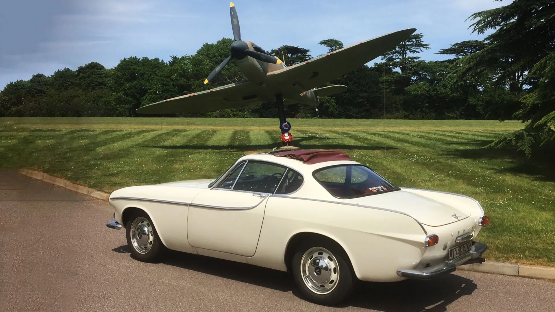 Rear side view of a Classic Volvo  P1800S in ivory with a burgundy webasto roof. Vehicle is parked in front of a military plane on display