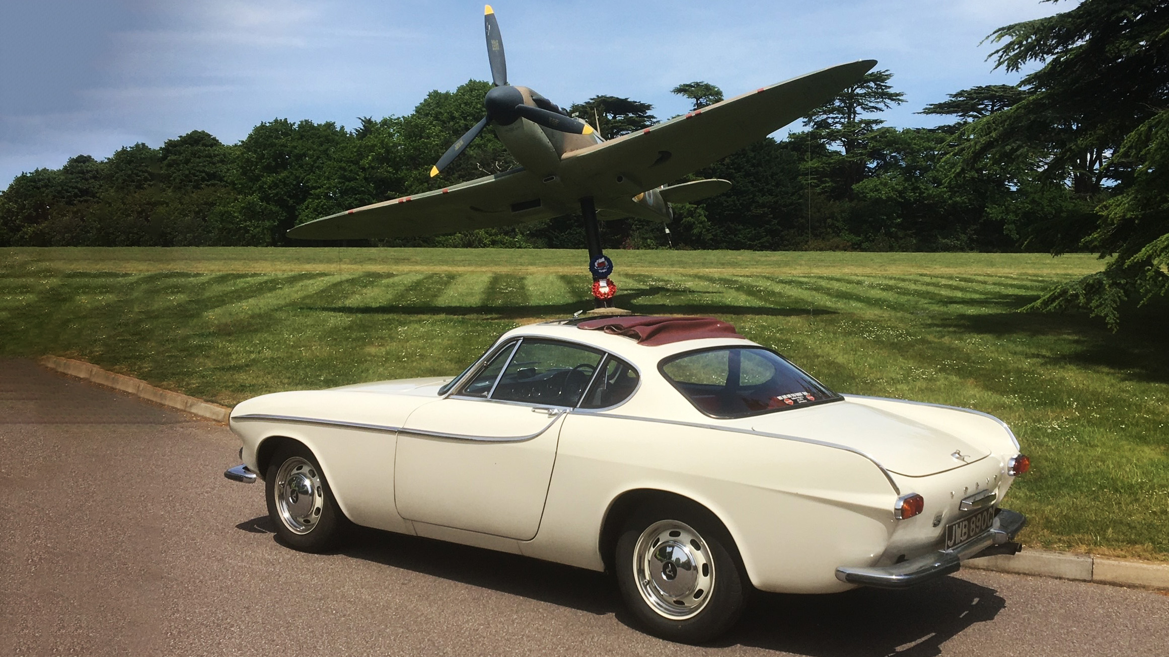 Rear side view of a Classic Volvo  P1800S in ivory with a burgundy webasto roof. Vehicle is parked in front of a military plane on display