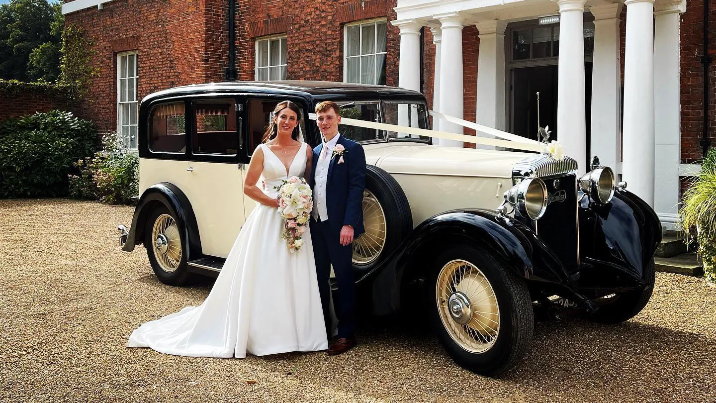 Vintage Daimler Limousine wedding car in Black and Ivory decorated with traditional white wedding ribbons and bow across its bonnet. Vehicle is parked in front of a local Batley we