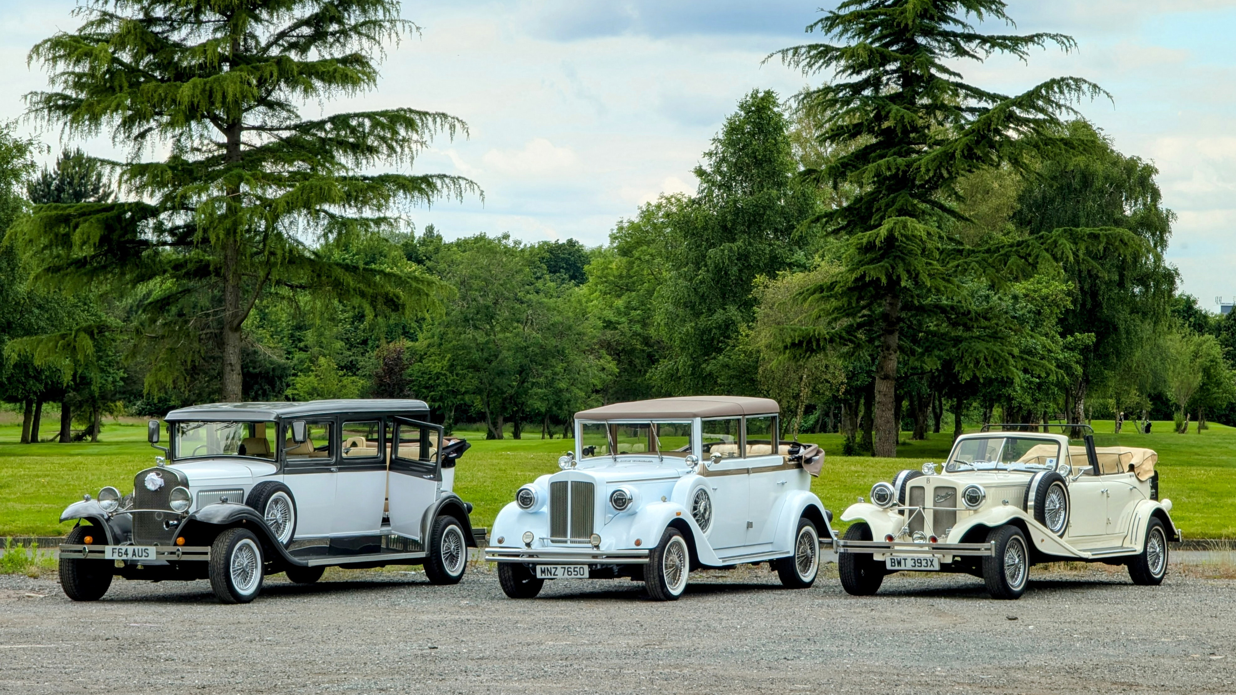 Three 1930s vintage Style convertible Wedding Cars. From left to right we have an ivory Bramwith, White Regent and an Ivory Beauford. Photo taken in a local West Yorkshire park with green trees in the background.