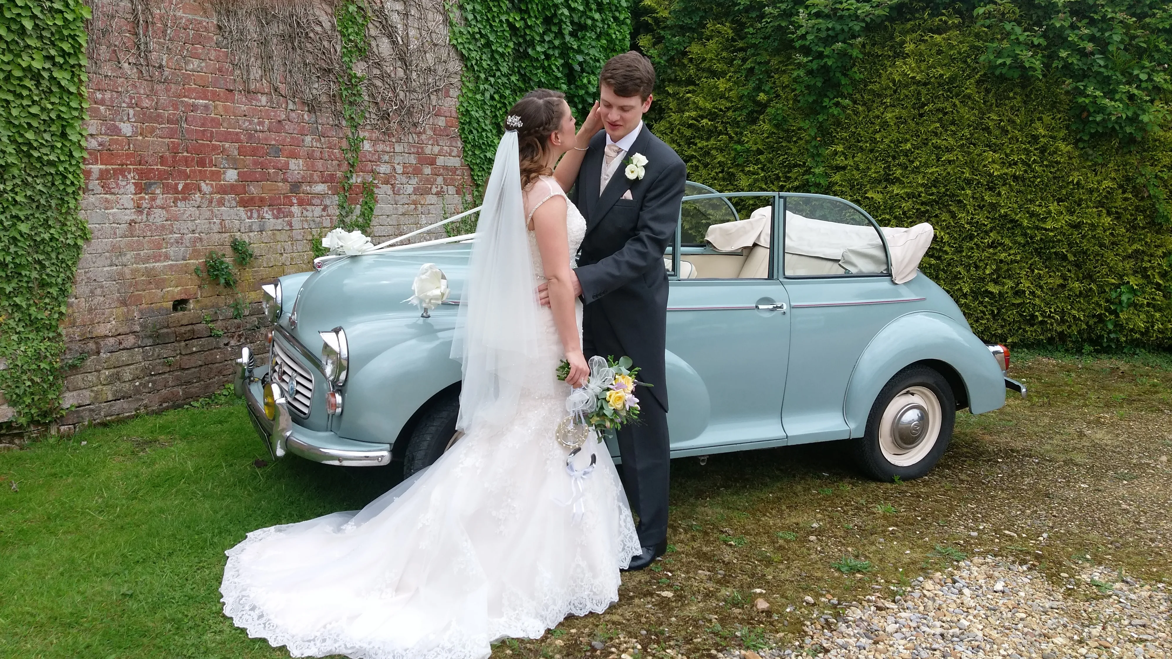 Andover-based Classic Morris Minor with ribbons and bow in light blue with its roof open showing a light cream interior. Bride and Groom are in front of the vehicle posing for their wedding photographer.