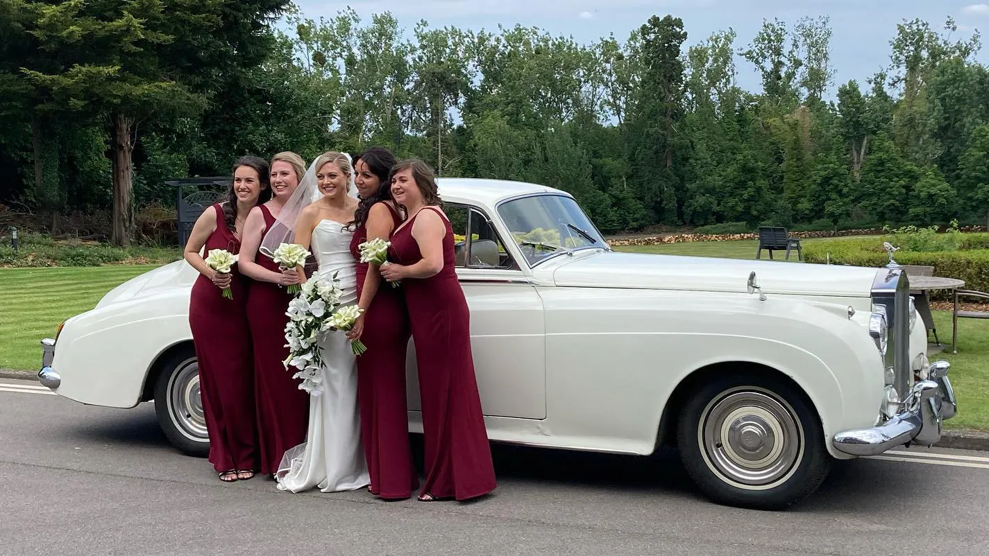 Classic white Rolls-Royce Silver Cloud parked  in front of a local Fleet wedding venue. Bride and four bridesmaids are standing in front of the vehicle for photos. The bride is wearing a white dress holding a large white bouquet of flowers. The bridesmaids are wearing Burgundy dresses with a smaller bouquet.